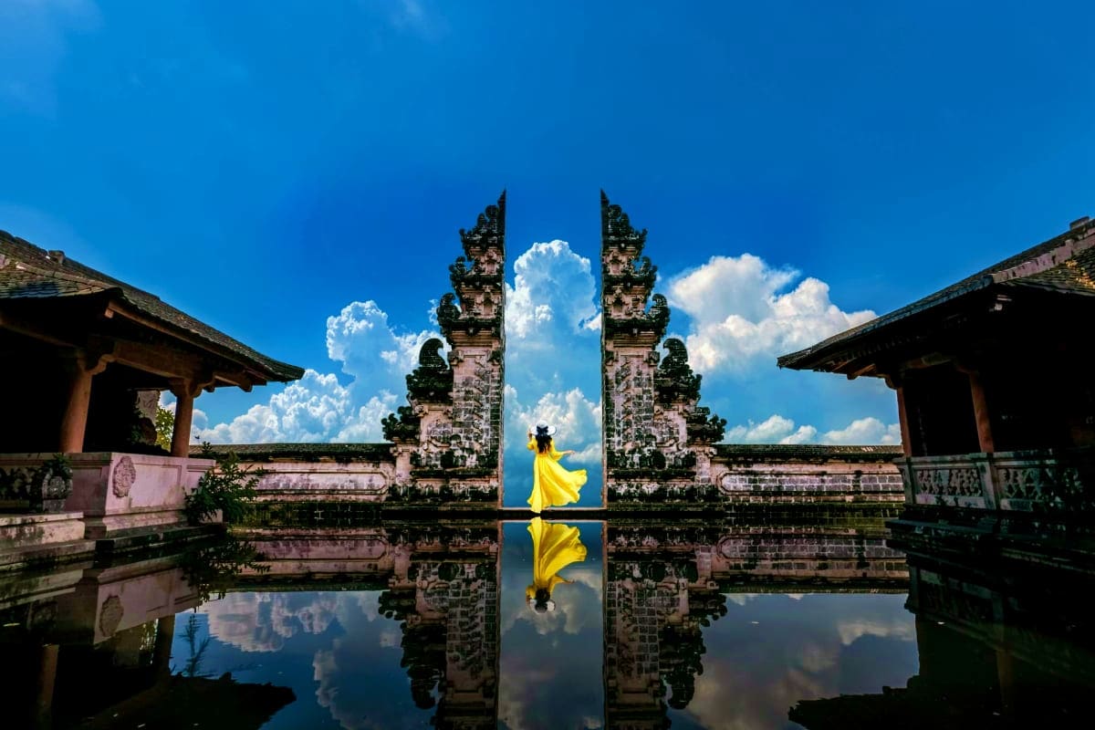Woman in yellow dress walking through ornate stone temple gates reflected in still water, Bali Indonesia