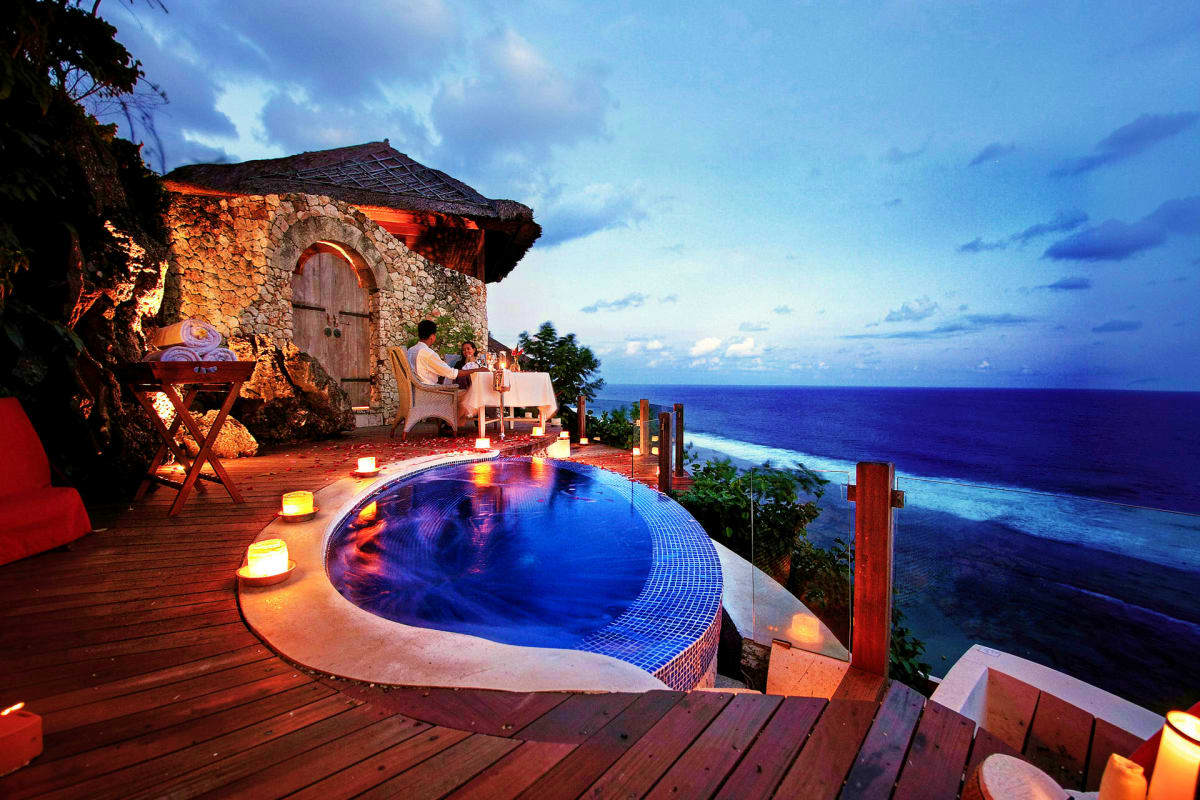 Luxury oceanfront villa in Bali with illuminated infinity pool overlooking the ocean, stone structure with arched doorway, wo
