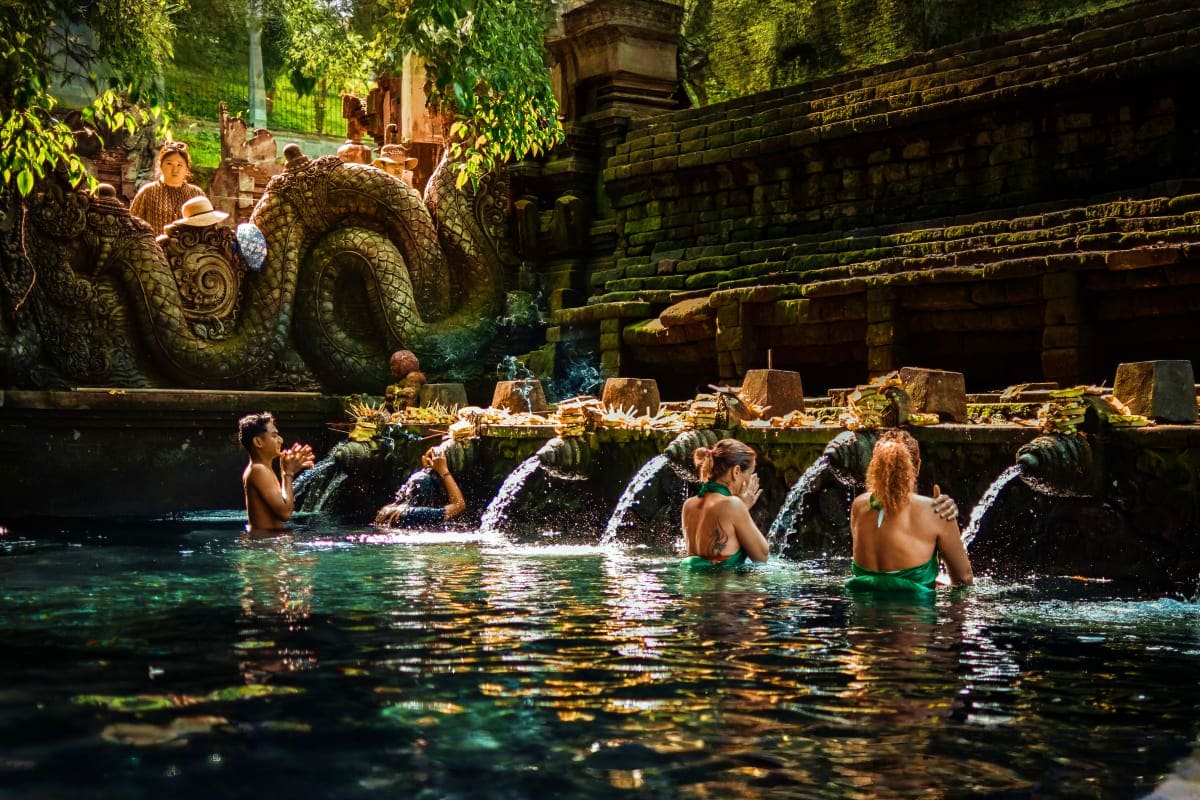 Tourists bathing in sacred spring water at Tirta Empul temple in Bali with ornate stone carvings and cascading water spouts