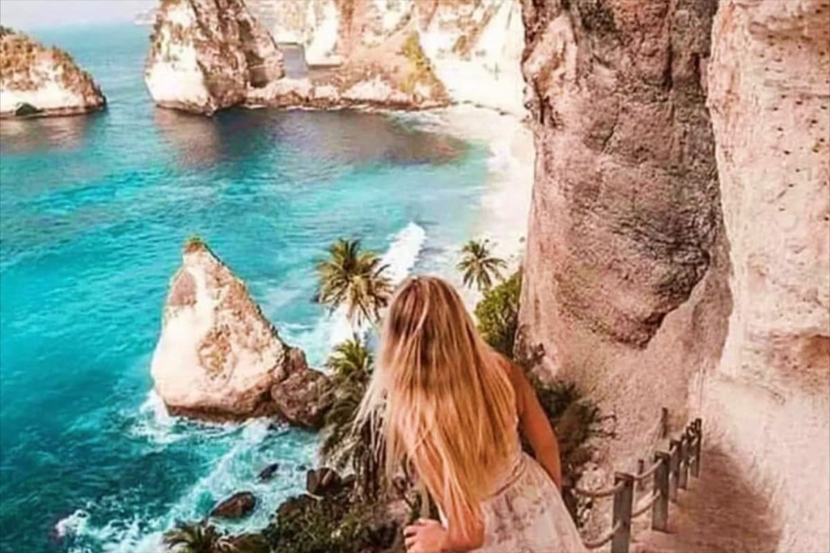 Woman with long hair standing on cliff edge overlooking turquoise ocean, white limestone rocks, and palm trees at tropical be