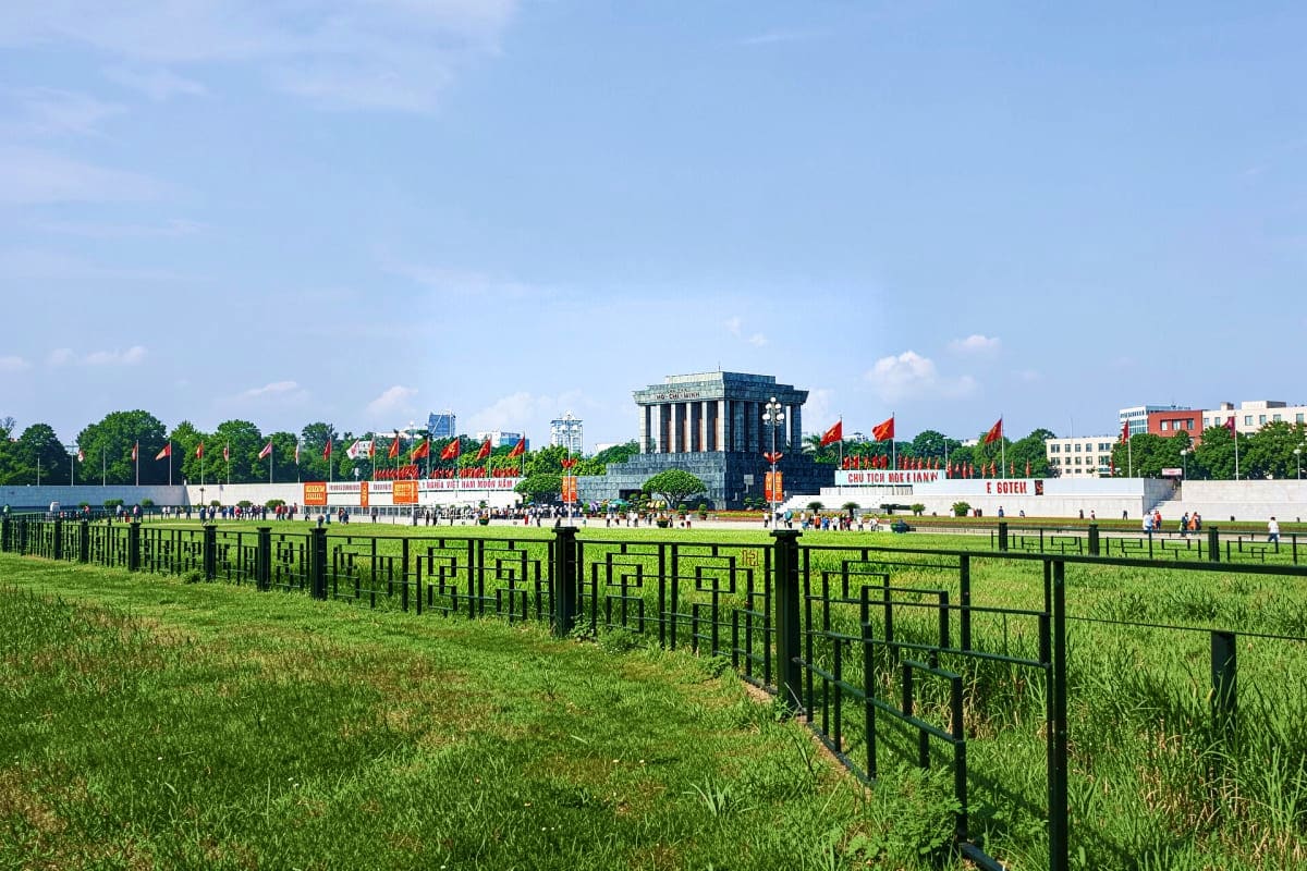 Ho Chi Minh Mausoleum in Hanoi with Vietnamese flags, green lawn, black fence, and crowds of visitors gathered in front of th