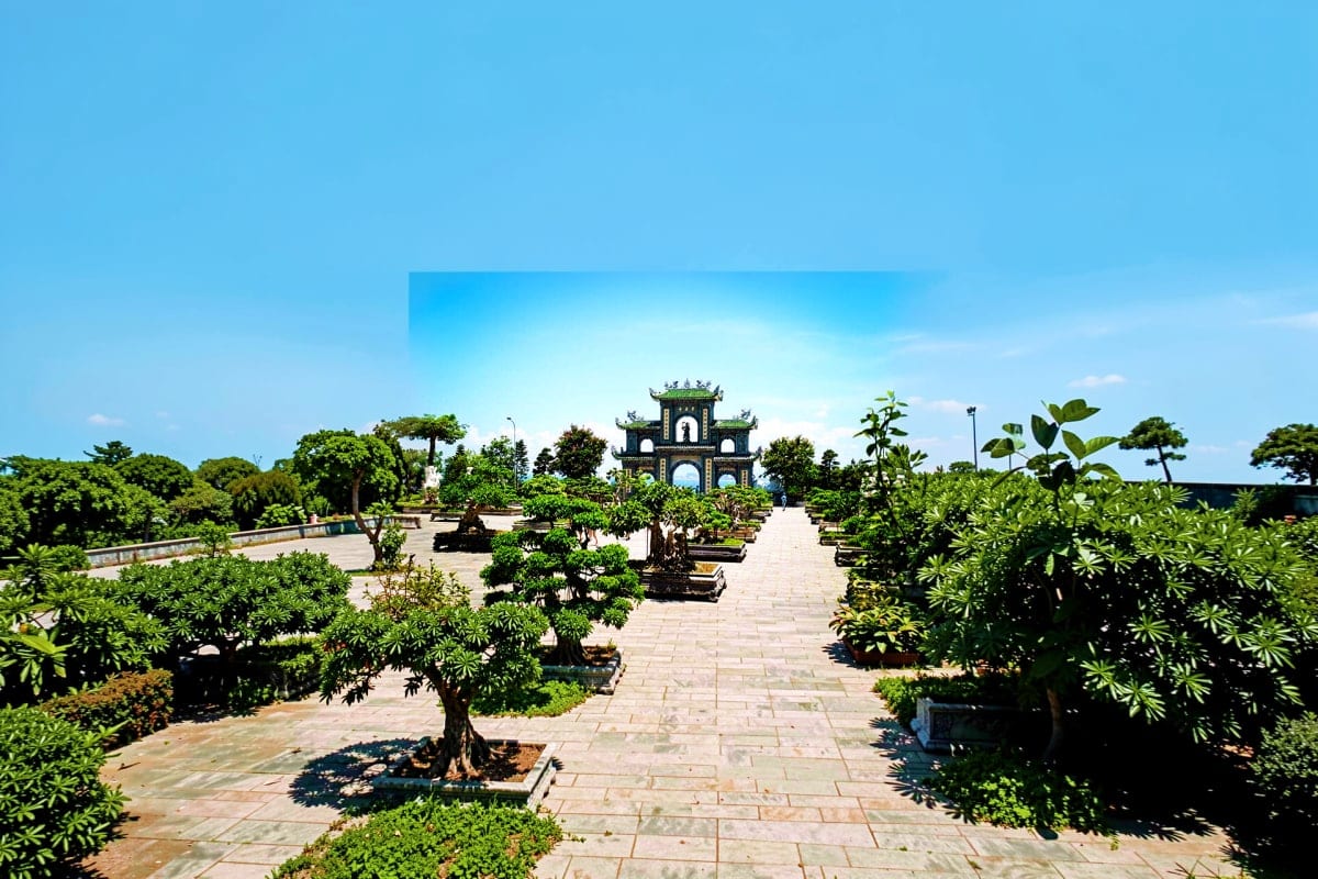 Historic Vietnamese temple with traditional architecture framed by manicured bonsai trees and stone pathway under clear blue 