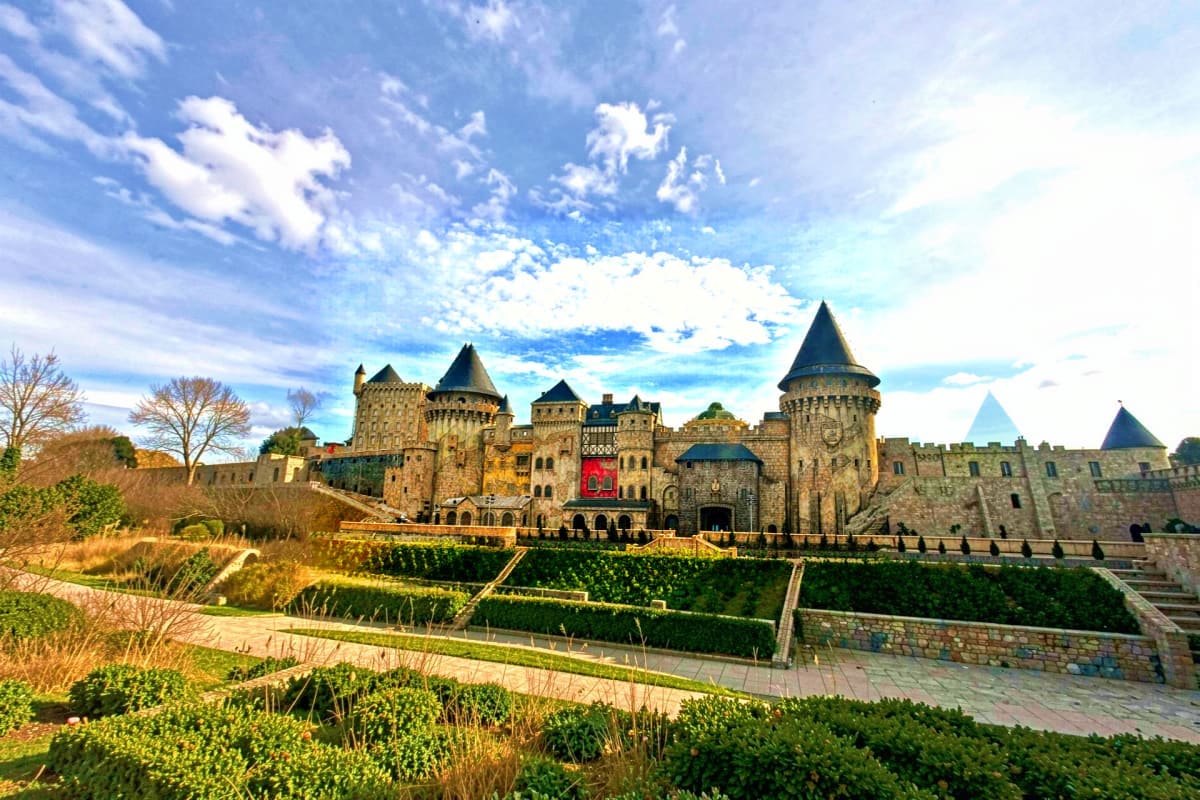 Medieval castle with stone towers and cone-shaped turrets surrounded by manicured gardens under blue sky with white clouds