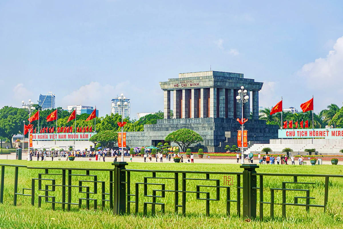 Ho Chi Minh Mausoleum in Hanoi with Vietnamese flags, green lawn, and visitors gathered at the iconic monument under blue sky