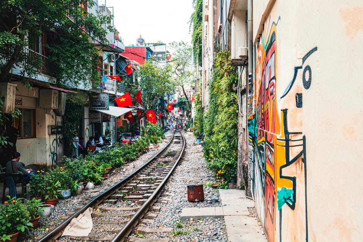 Narrow alley with railroad tracks, ivy-covered buildings, Vietnamese flags, and colorful street art in Hanoi's Old Quarter