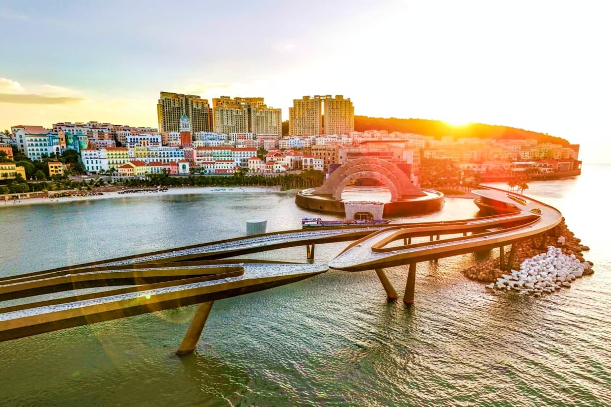 Coastal cityscape at sunset with curved pedestrian bridges, modern architecture, and a red circular building overlooking turq