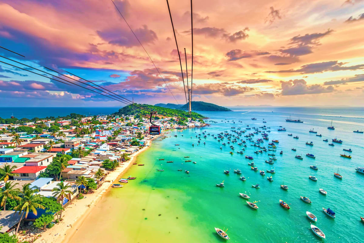Aerial view of Phu Quoc island with colorful fishing boats in turquoise waters, sandy beach, hillside village, and dramatic s