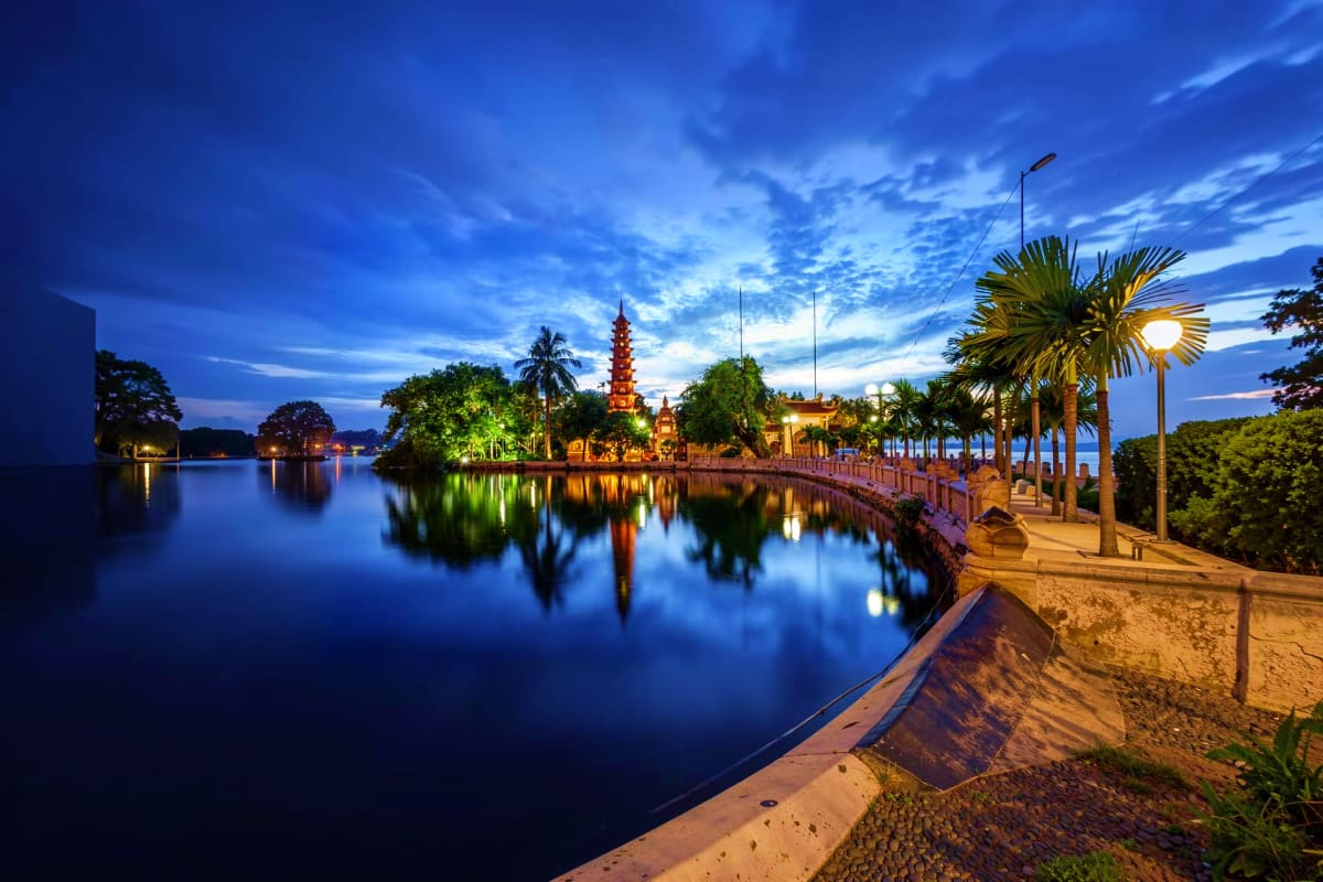 Tran Quoc Pagoda illuminated at twilight reflecting in Hoan Kiem Lake with palm-lined promenade in Hanoi, Vietnam