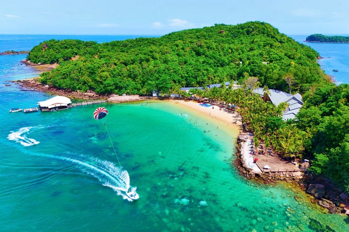 Aerial view of Phu Quoc beach with emerald water, parasailing activity, sandy shore, and lush green forested island landscape