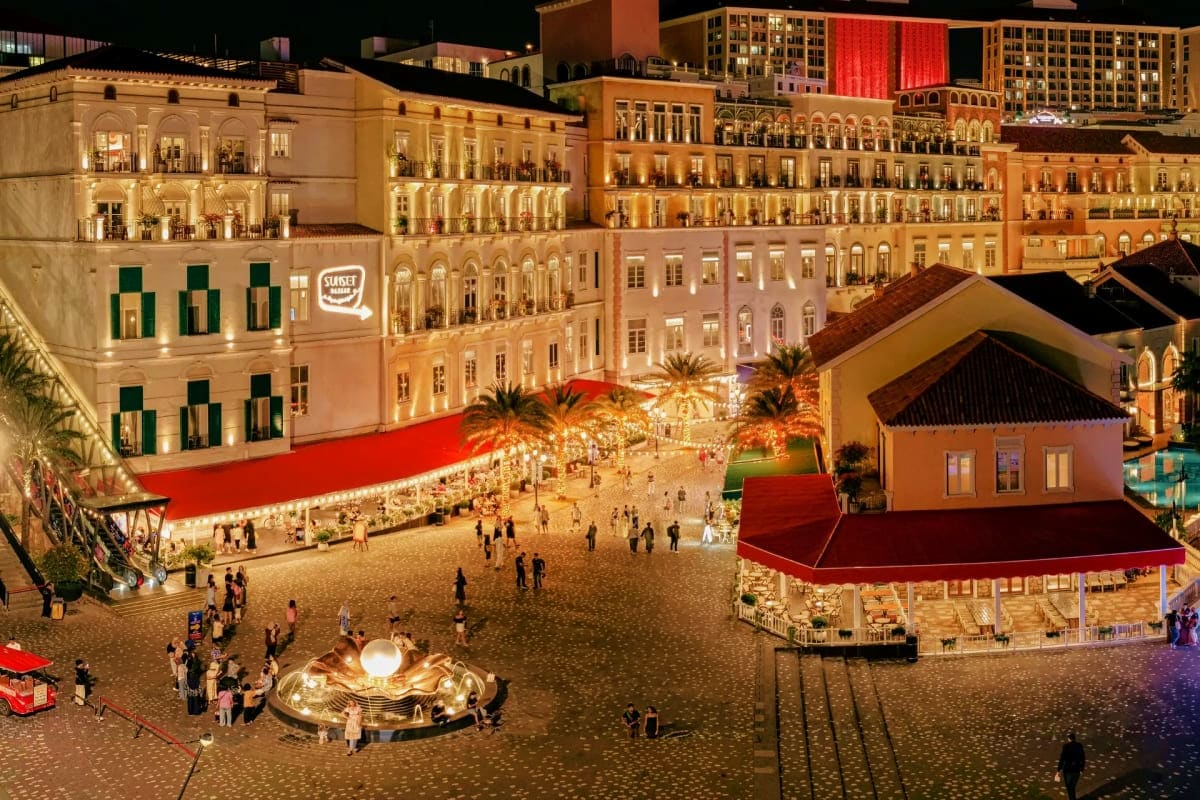 Illuminated seaside town square at night with colonial architecture, palm trees, red awnings, and visitors walking around a g