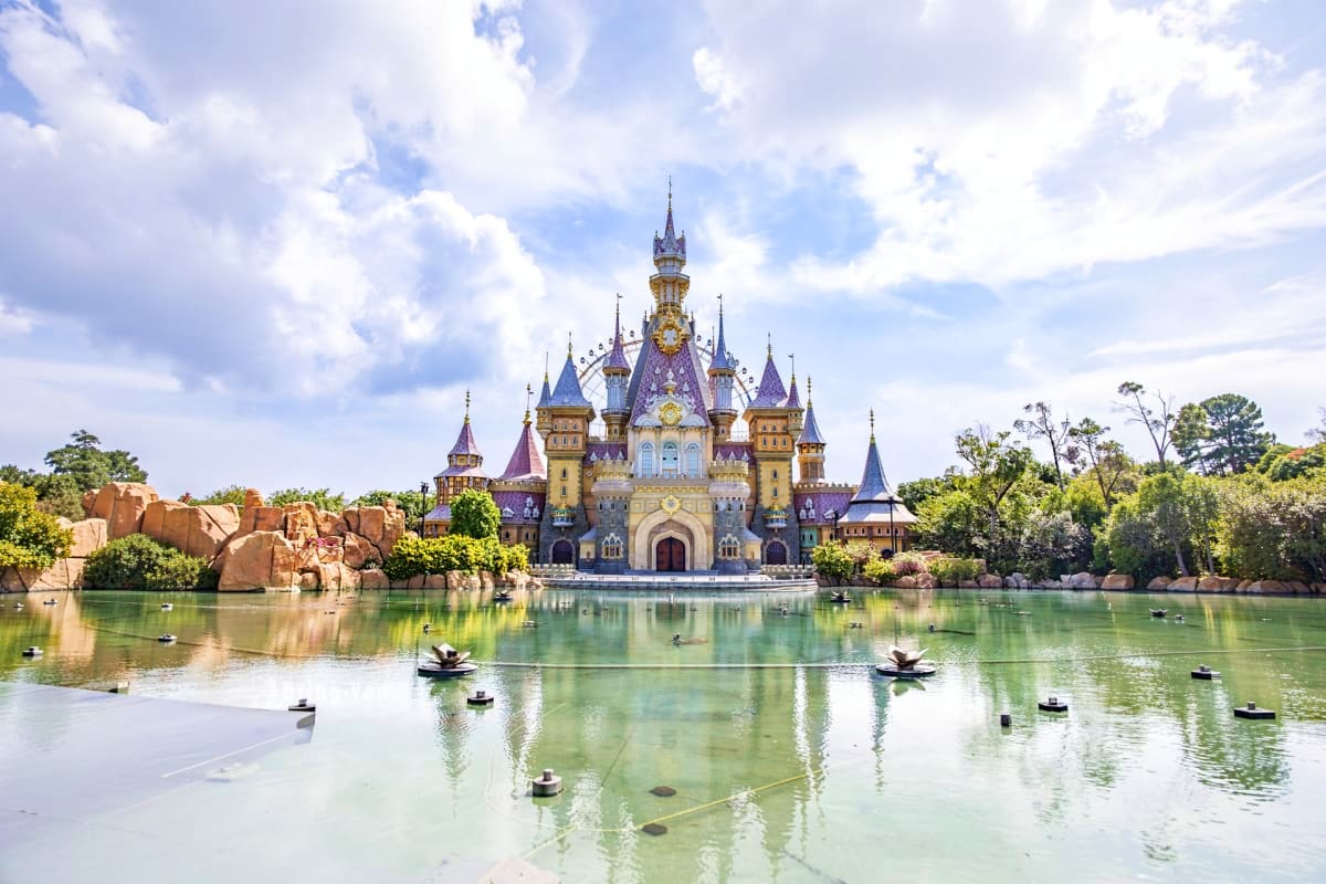 Ornate fairytale castle with purple and gold spires reflected in calm green water, surrounded by lush trees and rocky outcrop