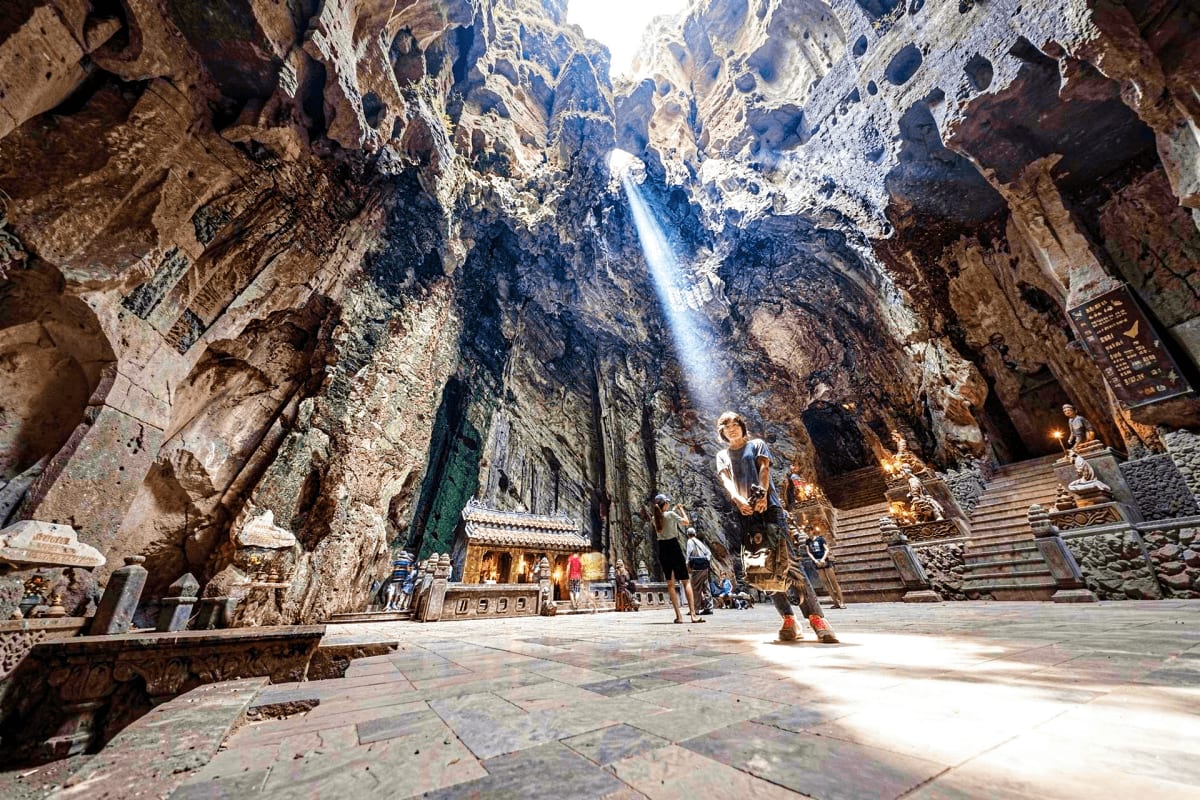 Sunlight streams into massive cave sanctuary with ornate Buddhist temple, golden statues, stone staircase, and tourists explo