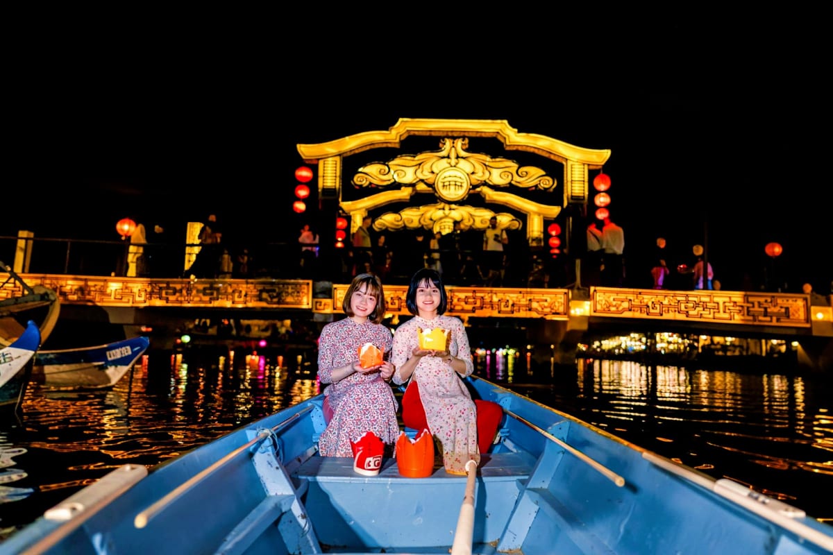 Two women in traditional áo dài dresses floating on a wooden boat at night with illuminated golden Chinese gate and red lante