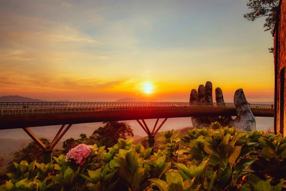 Golden Gate Bridge at sunset with giant stone hand sculpture and pink flowers in foreground, Vietnam landscape
