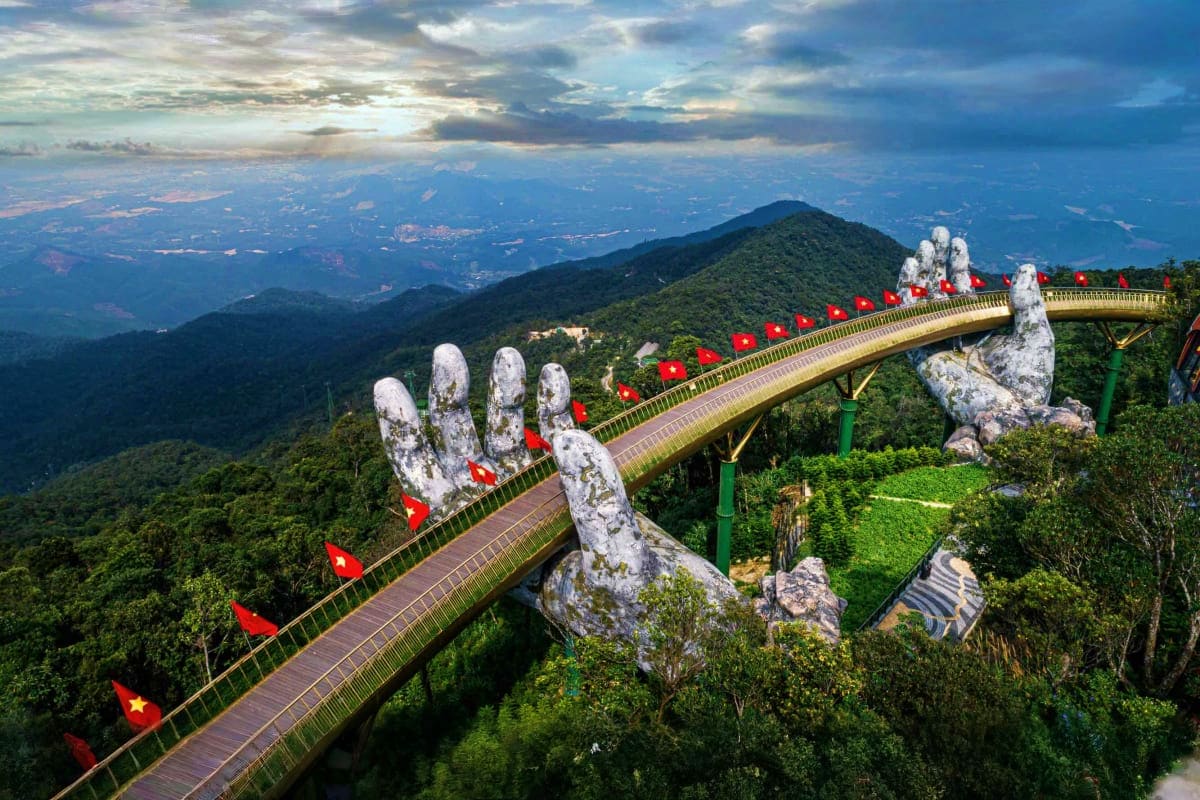 Golden Bridge with giant stone hands supported by green structure in mountainous Vietnamese landscape with red flags and fore