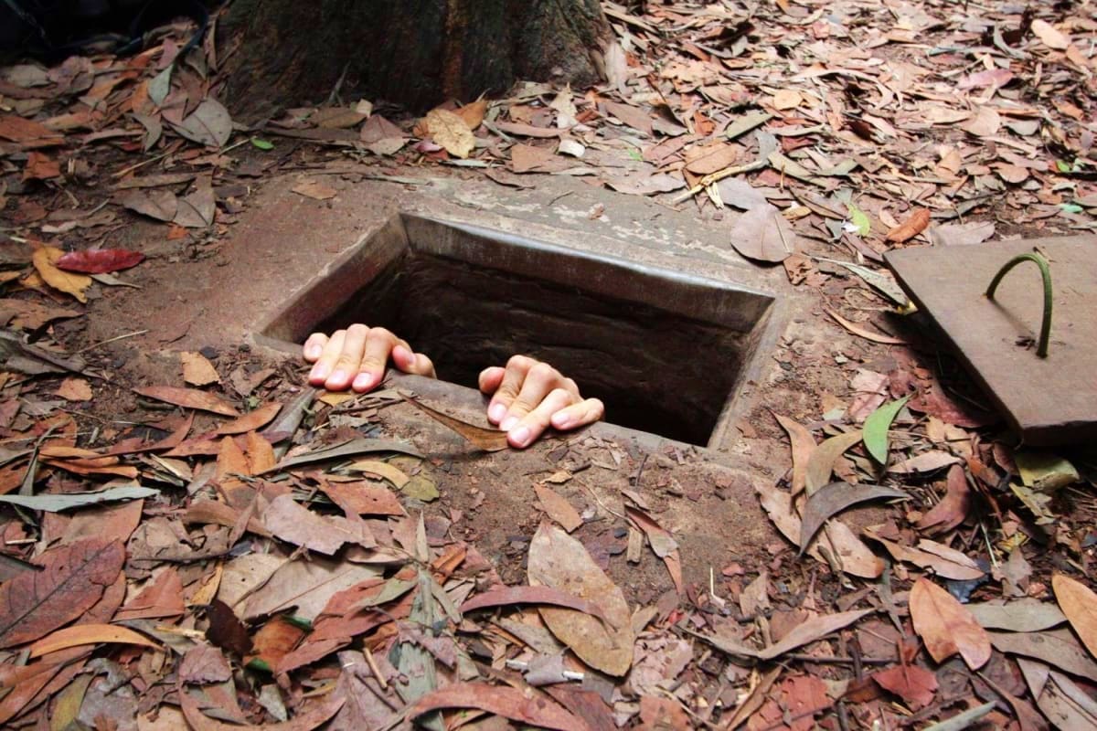 Hands emerging from underground bunker tunnel surrounded by fallen leaves and forest debris at Cu Chi Tunnels