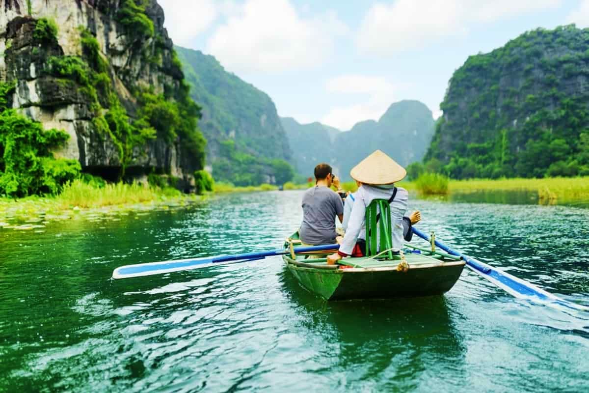 Two tourists rowing a traditional wooden boat through Ninh Binh's limestone karst mountains and emerald water in Vietnam