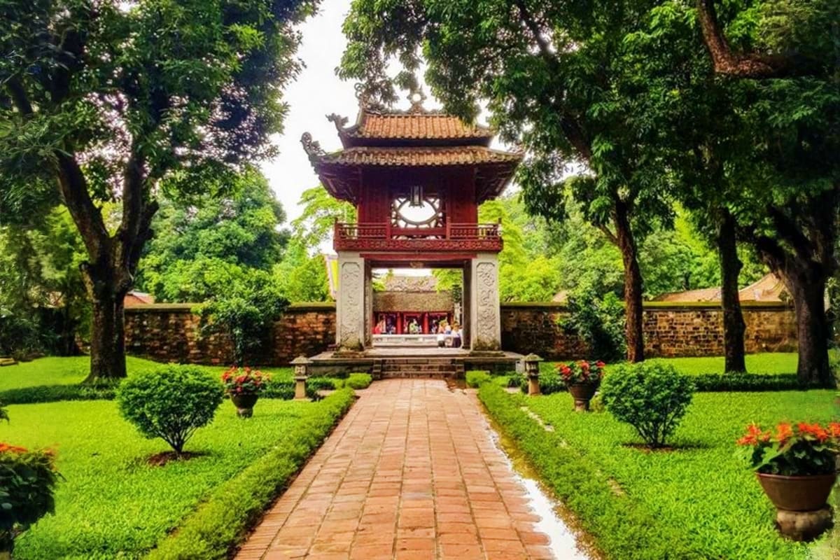 Traditional Vietnamese temple with red-tiled roof and clock tower framed by lush green trees and manicured gardens along bric