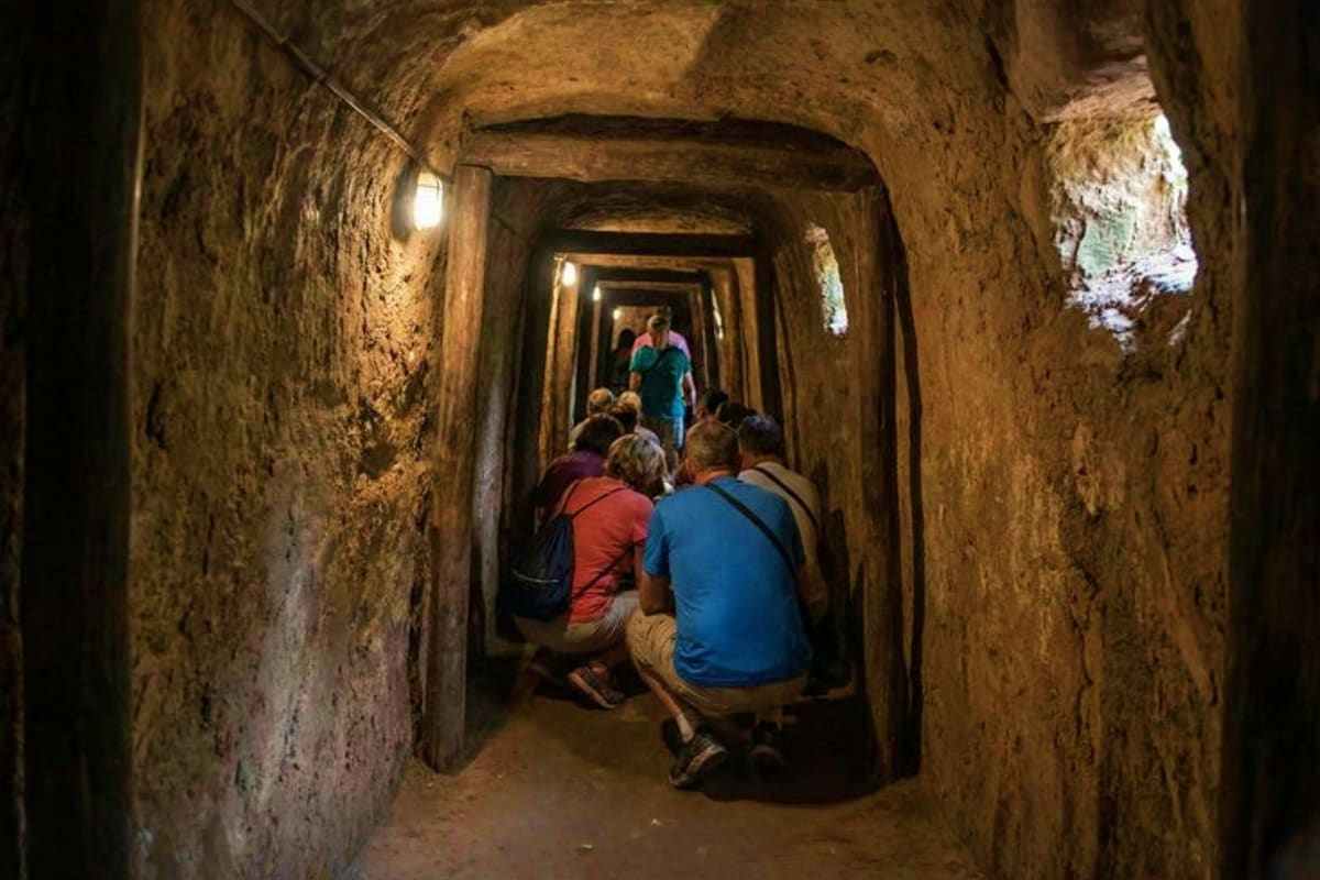 Tourists exploring a narrow underground tunnel with arched stone ceiling and dim lighting, part of historical war tunnels in 
