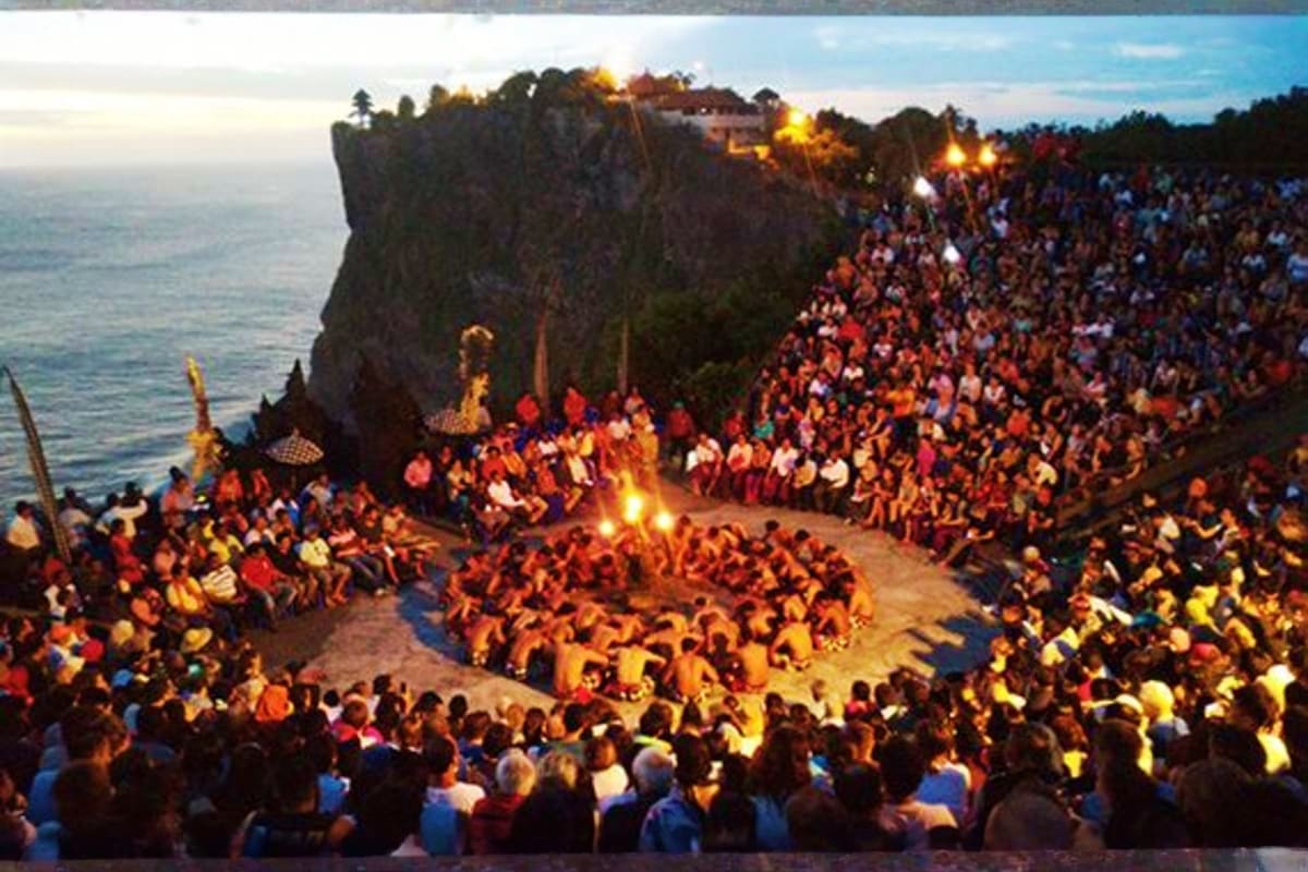 Large crowd gathered on beach at clifftop temple during evening fire ceremony ritual in Bali, Indonesia.