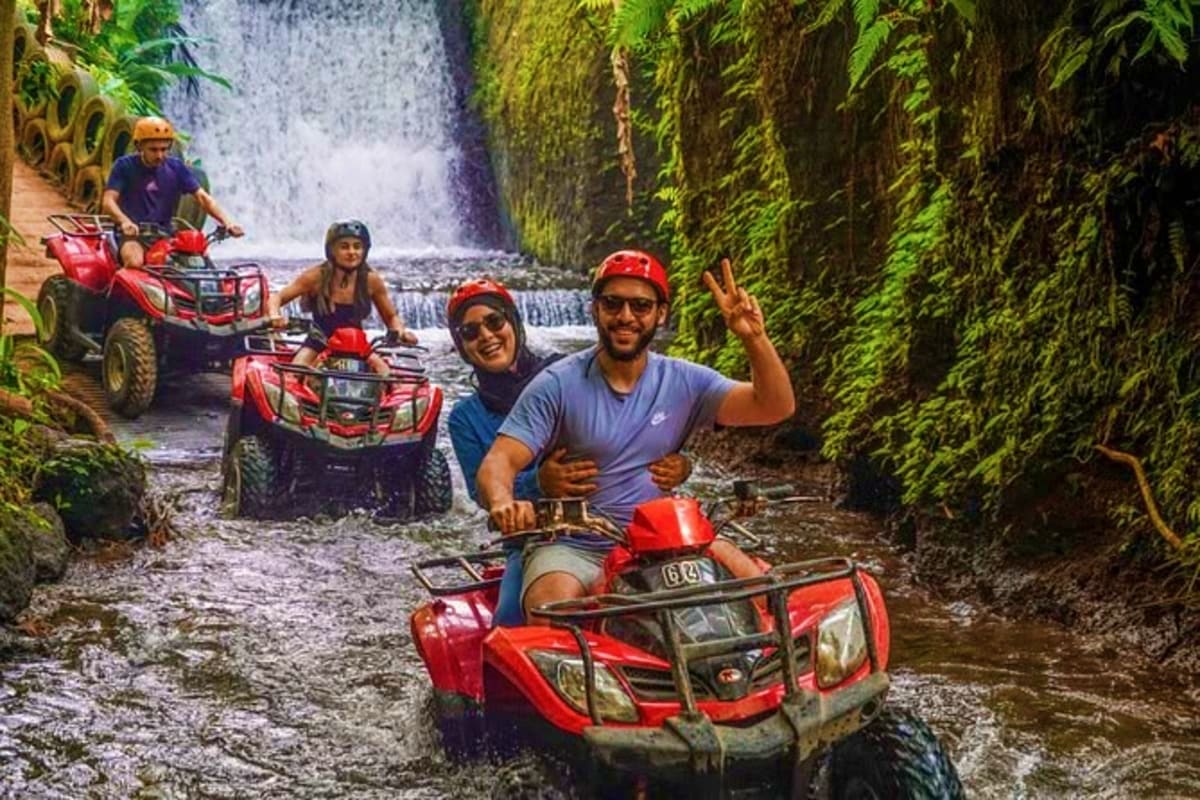 Group of tourists on red ATVs riding through lush jungle canyon with waterfall in background during Bali adventure tour