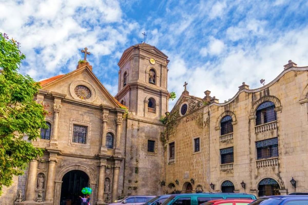 Best of Manila and Boracay Holiday Package Historic stone cathedral with bell tower and cross in Manila plaza, featuring Spanish colonial architecture and parked vehicl