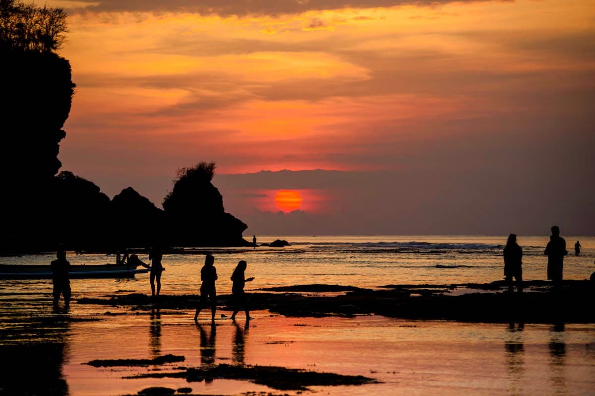 Silhouetted figures wading and surfing at sunset on a tropical beach with rocky cliffs and golden sky reflecting on the water