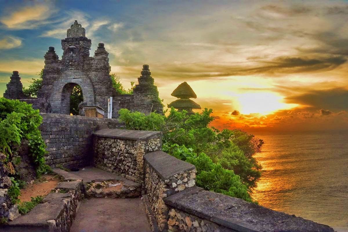 Ancient stone temple ruins overlooking golden sunset over ocean water with lush green vegetation on coastal cliffs in Bali