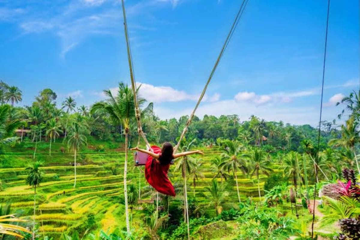 Woman in red dress on rope swing above lush green rice fields and palm trees in Bali
