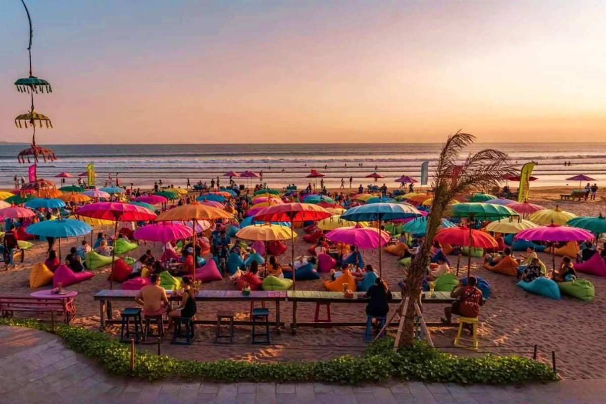Crowded beach at sunset with colorful umbrellas, bean bag seating, and tourists enjoying the sandy shore in Bali