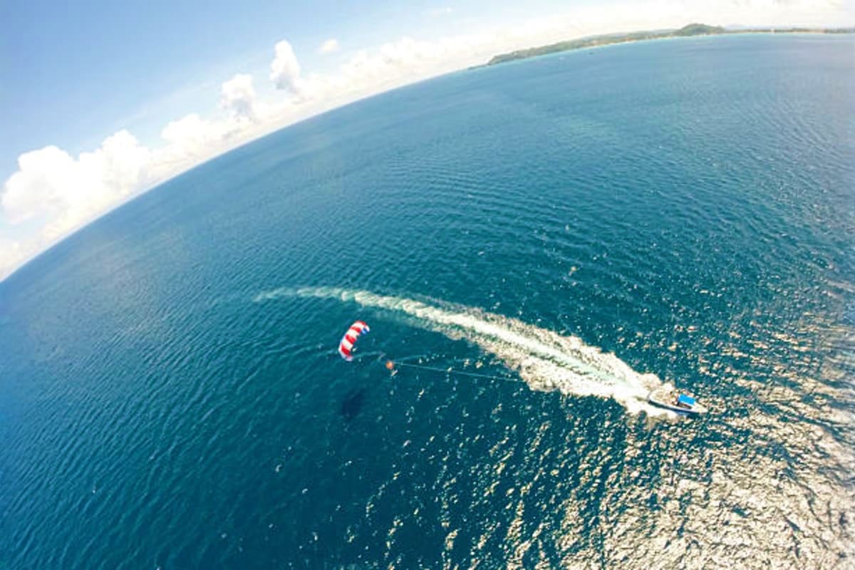 Aerial view of a parasailer with red and white parachute towed behind a speedboat in turquoise ocean water near Bali coastlin