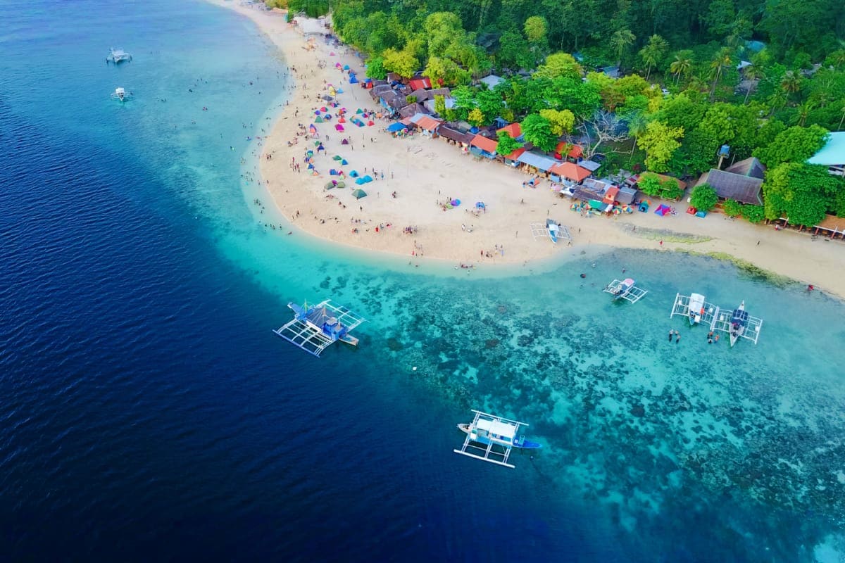 6 Days Manila and Cebu Holiday Package Aerial view of tropical beach with white sand, turquoise water, traditional outrigger boats, and dense green jungle vegetatio