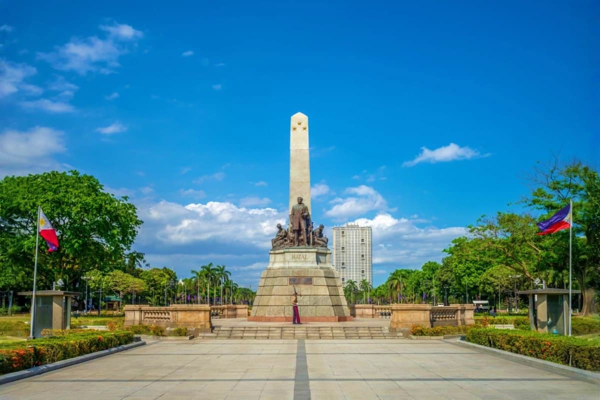 Best of Manila and Boracay Tour Package Rizal Monument in Manila with tall obelisk, bronze statues, palm trees, and Philippine flags under blue sky