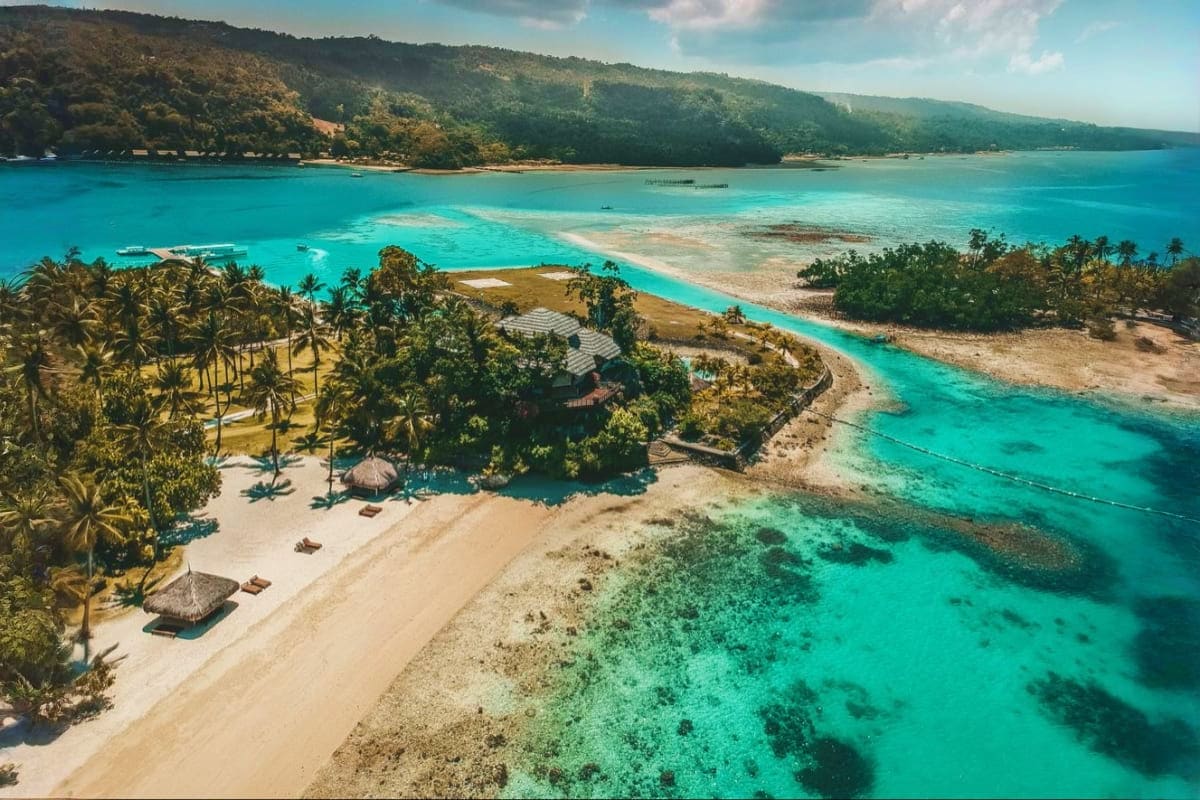 Davao to Siargao Adventure Tour Aerial view of tropical beach with turquoise waters, white sand, palm trees, and forested hills in Siargao Island