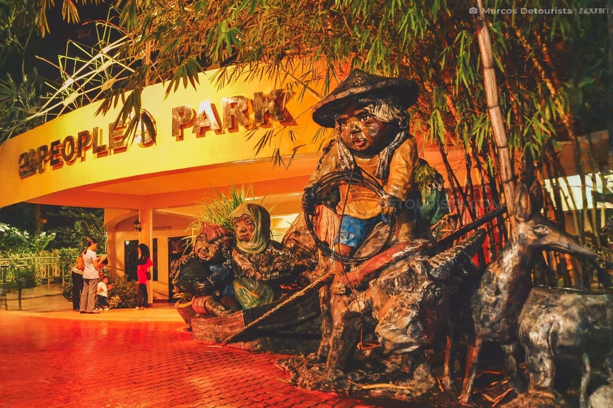 Davao to Siargao Adventure Tour People's Park entrance with bronze statues of indigenous figures and bamboo vegetation at night, Davao City.