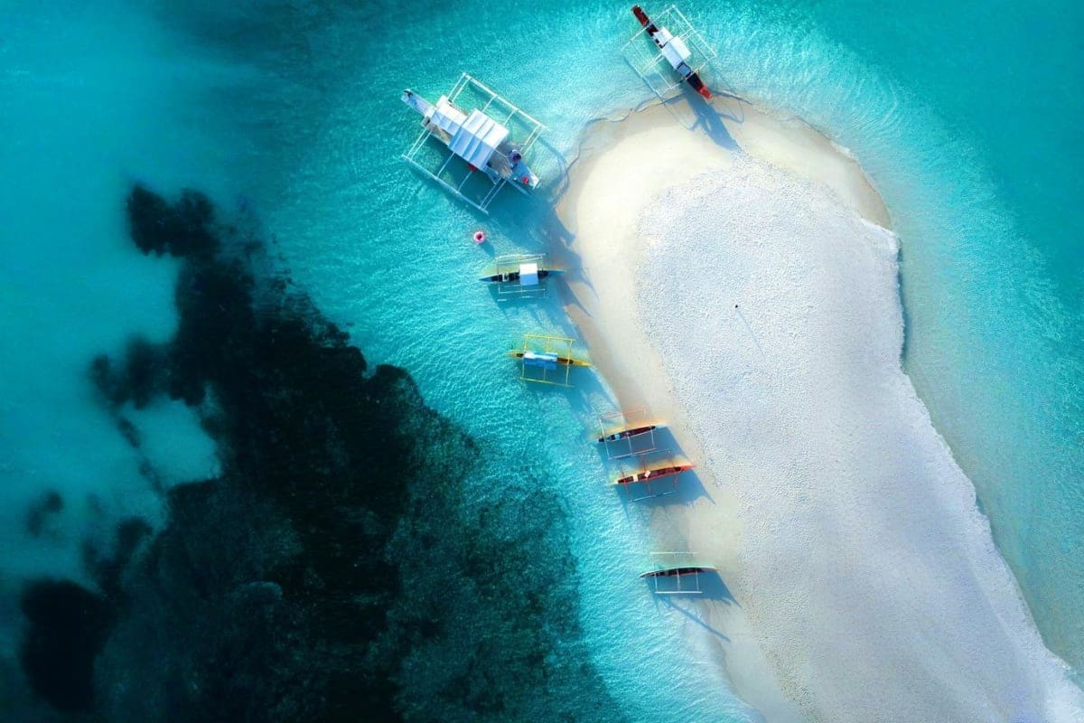 Davao to Siargao Adventure Tour Aerial view of white sand island with turquoise water, traditional boats moored offshore, and coral reef visible in shallow w
