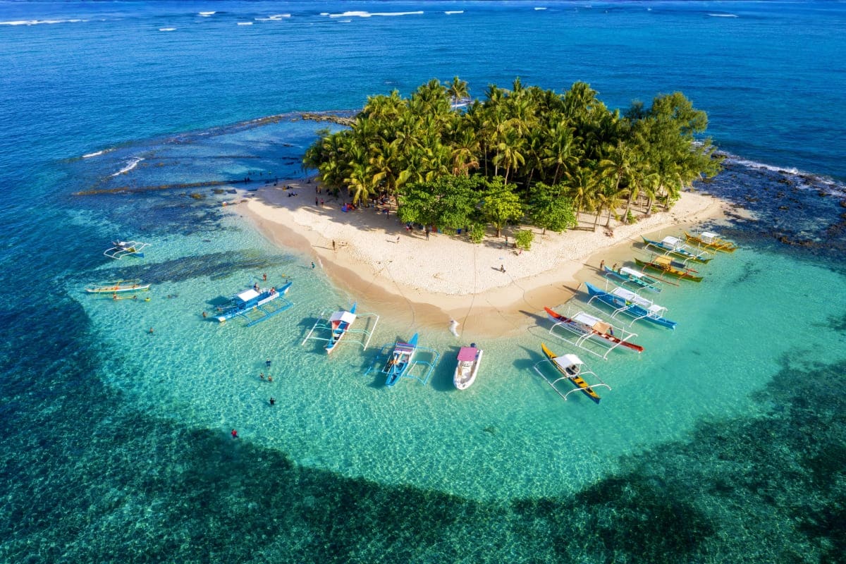 Davao to Siargao Adventure Tour Aerial view of a small tropical island with palm trees, white sand beach, and colorful outrigger boats moored in turquoise wa