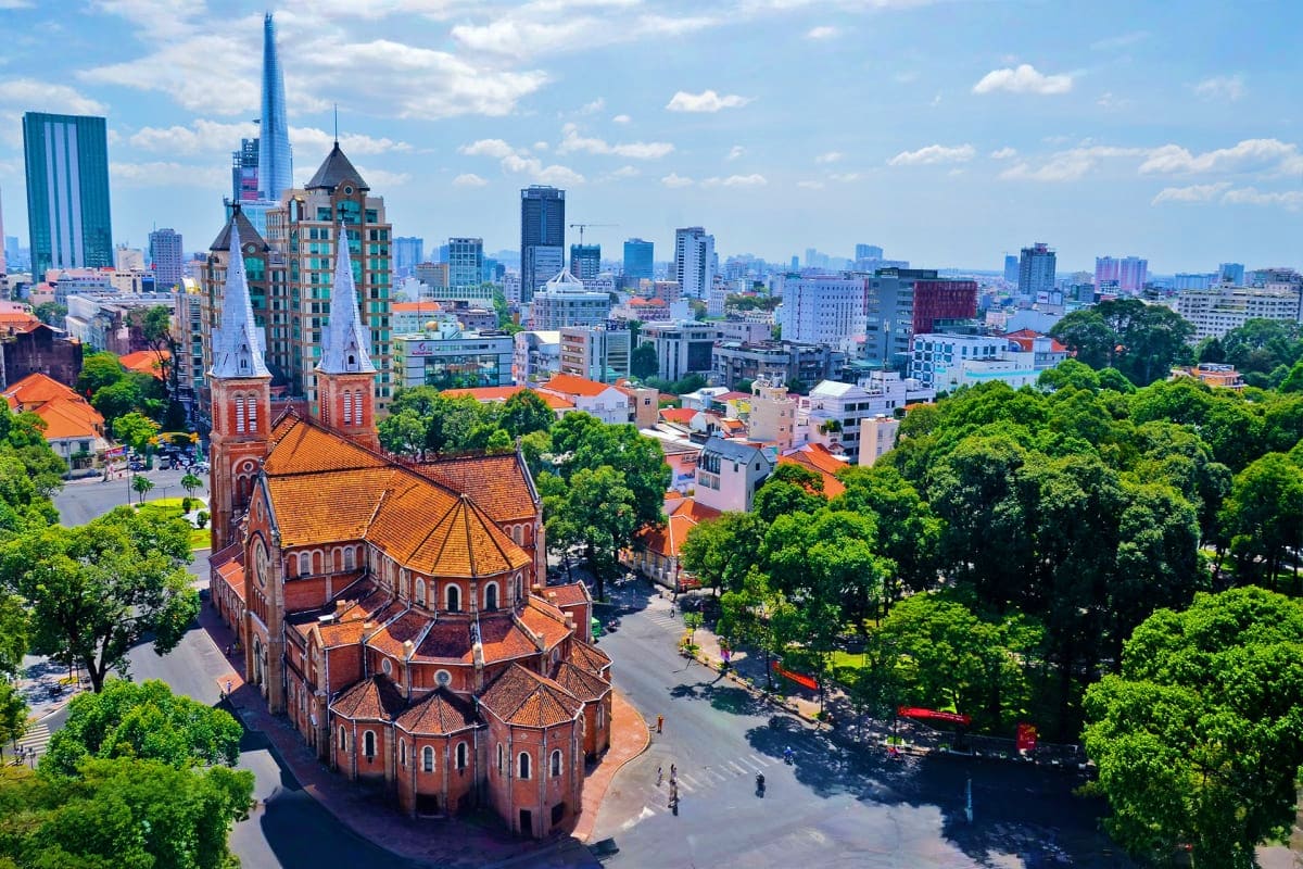 Aerial view of Saigon Notre-Dame Cathedral with red brick architecture surrounded by green trees and Ho Chi Minh City skyline