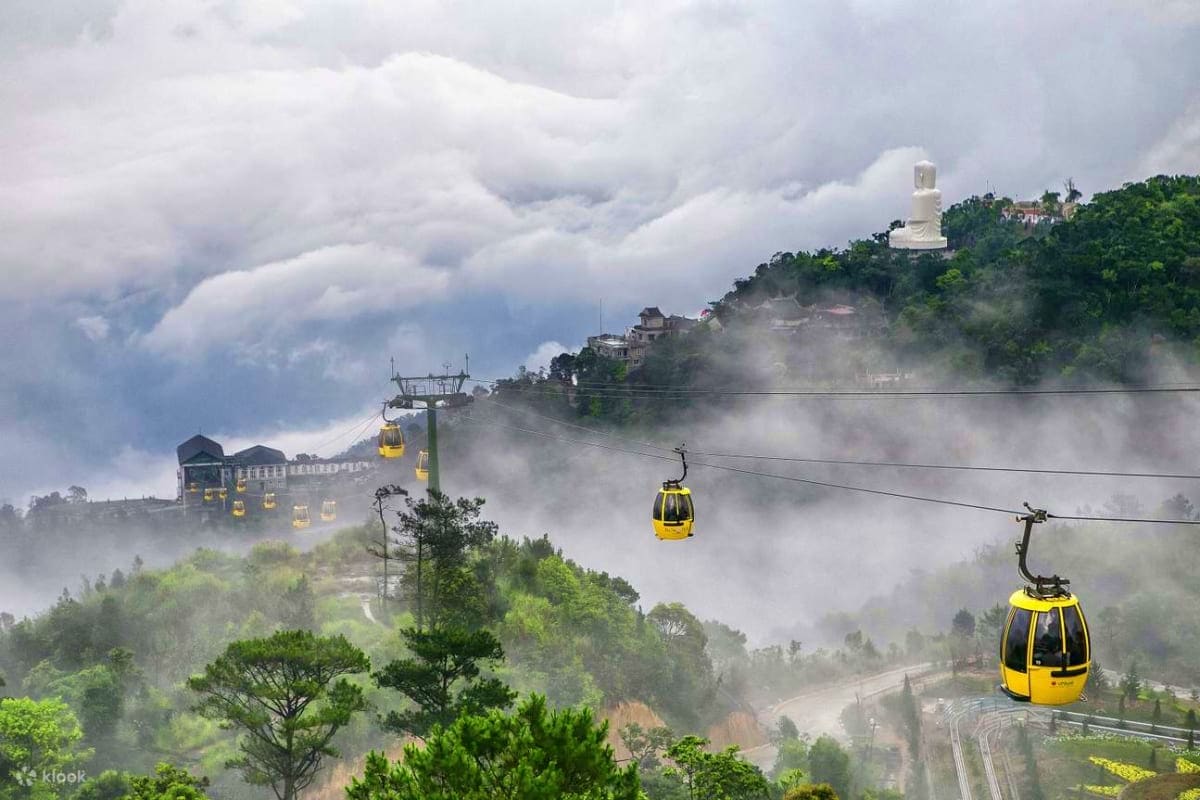 Yellow cable cars suspended over misty forested mountain landscape with temple buildings and white lighthouse visible on hill
