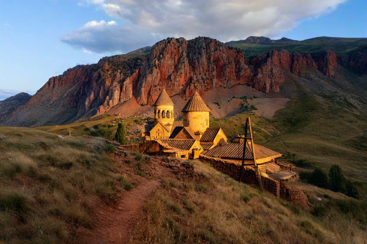 Yellow Armenian church with two domes nestled in mountainous landscape with red rocky cliffs and sparse vegetation