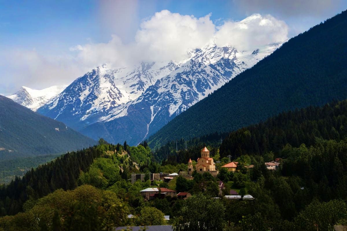Snow-capped mountain peaks overlooking a green forested valley with a traditional Orthodox church nestled among pine trees in