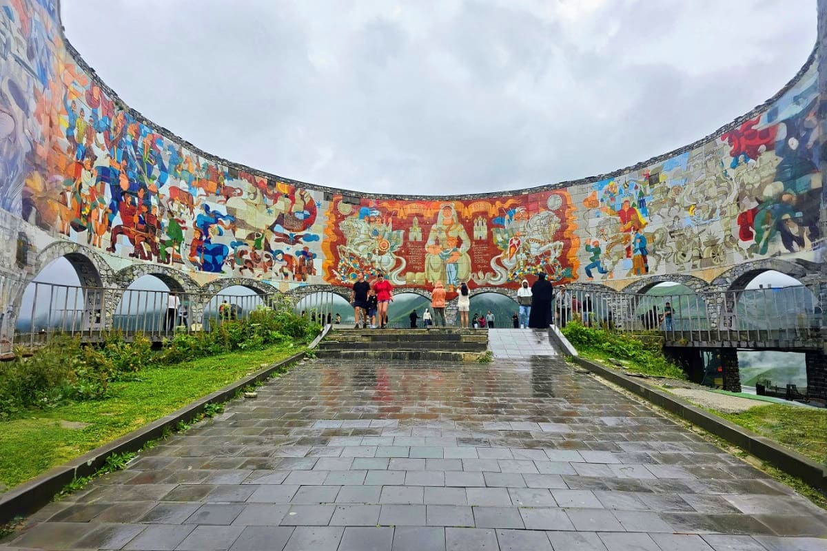 Circular courtyard with colorful murals depicting Georgian cultural scenes, stone arches, wet paved floor, and visitors explo