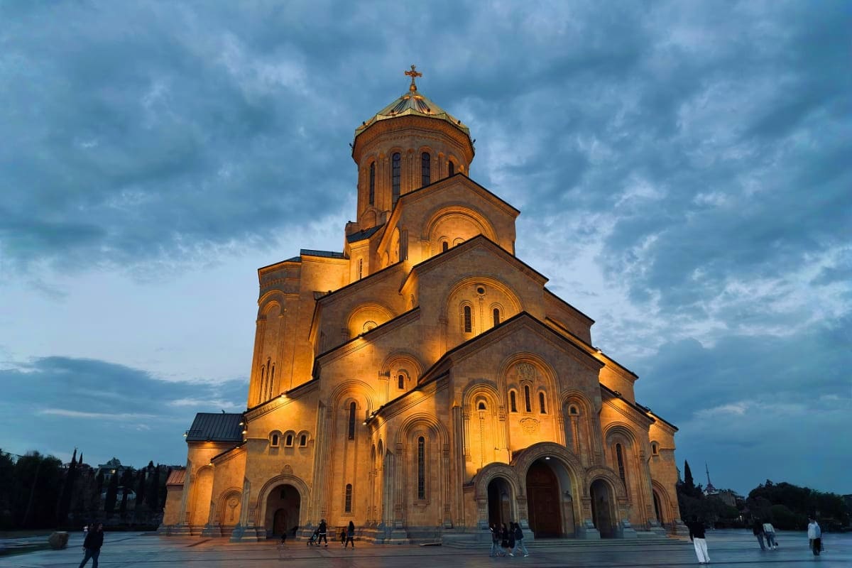 Orthodox cathedral with golden-lit domes and cross at dusk, stone facade, people in plaza, cloudy sky