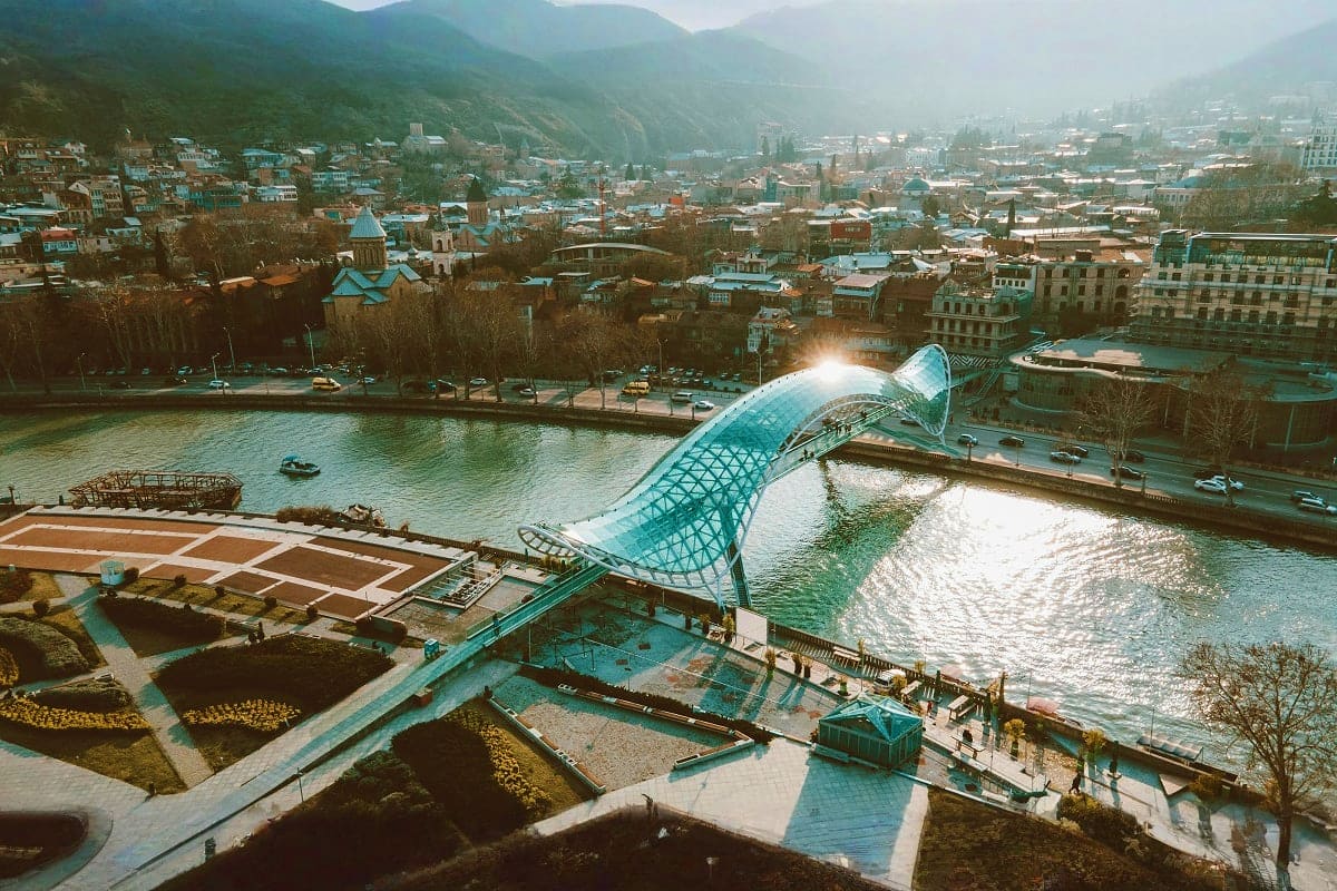 Aerial view of Tbilisi with modern glass bridge over Mtkvari River, mountains, and cityscape with historic buildings and chur