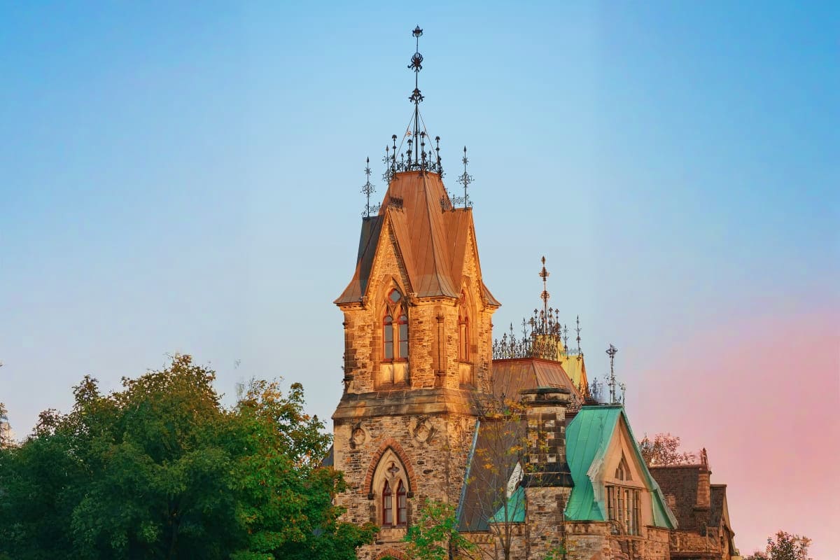 Gothic Revival castle tower with ornate spires and copper roof at sunset, surrounded by green trees under clear sky