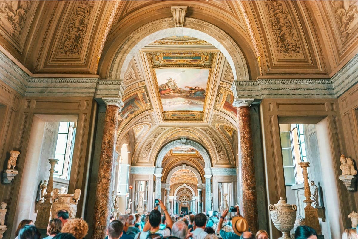 Ornate museum gallery with decorated arches, frescoed ceiling, marble columns, classical sculptures on pedestals, and tourist