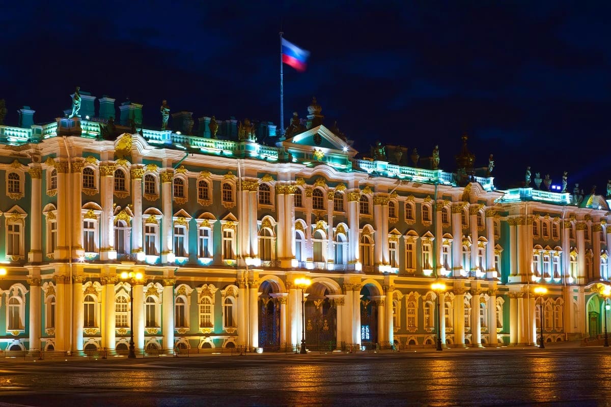 Winter Palace illuminated at night with Russian flag, reflecting in wet plaza during Moscow Imperial Journey tour
