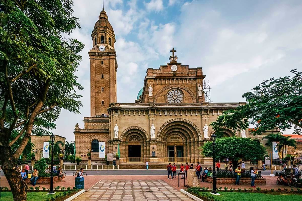 Historic stone cathedral with tall bell tower and cross, surrounded by green trees in Manila plaza with visitors walking