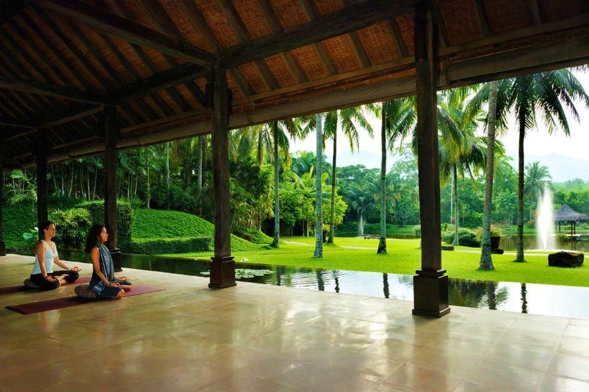 Two women sitting on yoga mats on a covered pavilion overlooking a manicured tropical garden with palm trees and a fountain