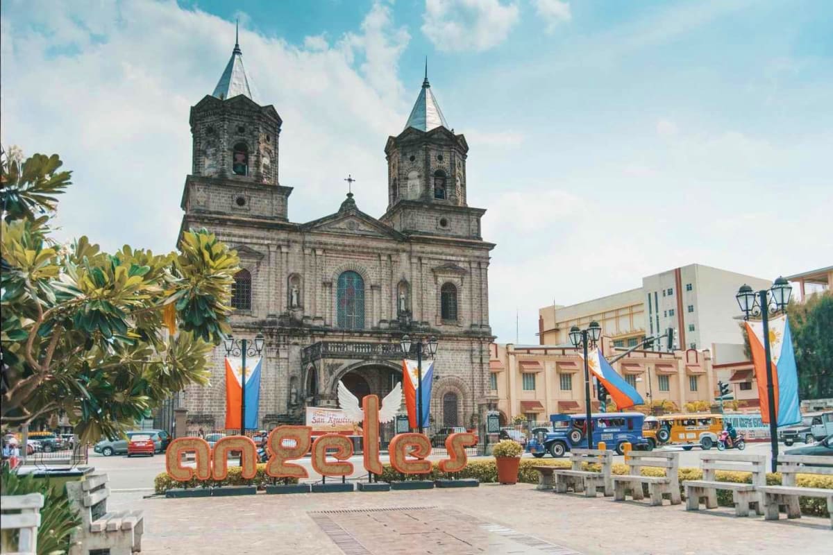 Manila Angeles Budget Tour Historic stone church with twin towers and cross, decorated plaza with "Angeles" sign and colorful banners, colonial architec